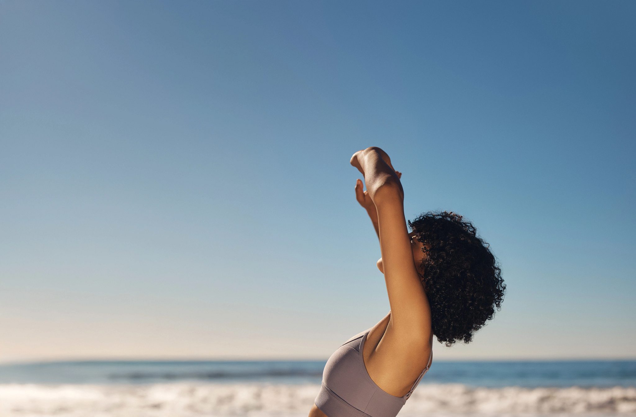An adult female stretching her arms above head while standing on a sandy beach with ocean in the background