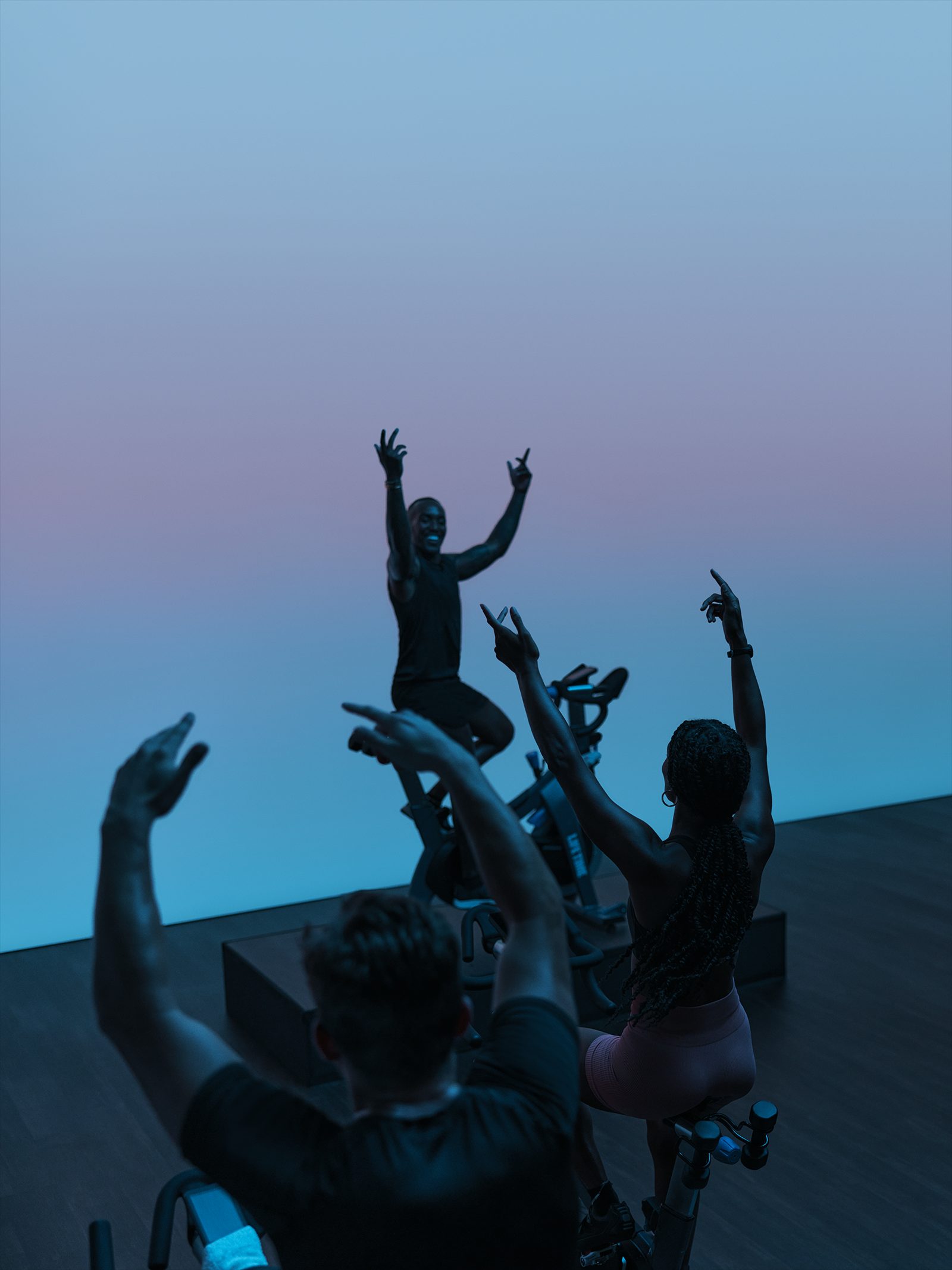 AMP spin class riding together, led by their instructor, with their hands in the air