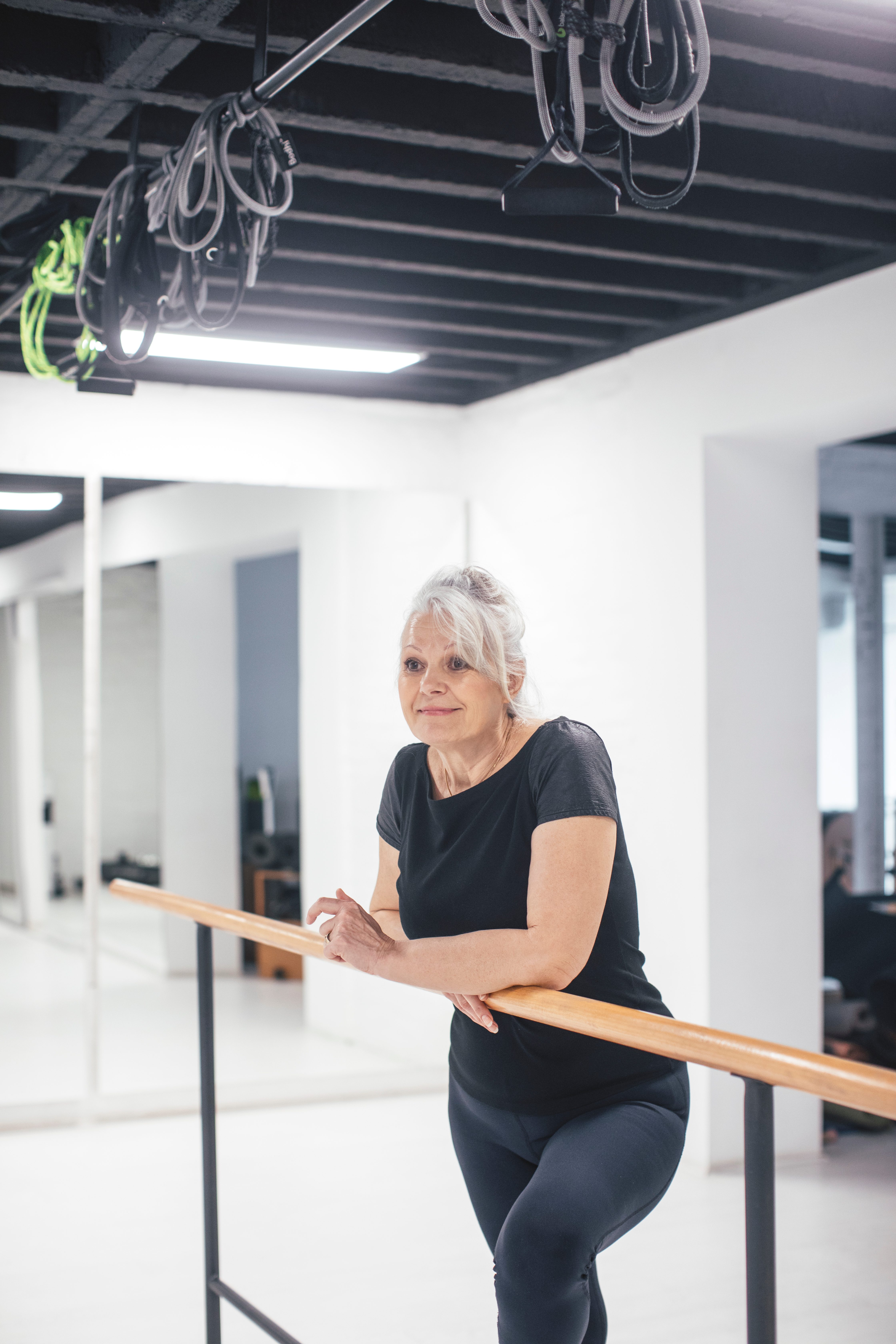 Pretty senior Caucasian woman standing by the ballet barre at gym.