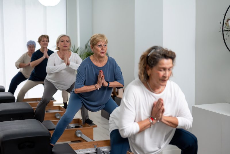 Group of concentrated mature women in sportswear performing side splits exercise on reformer machine while practicing Pilates in modern light studio.