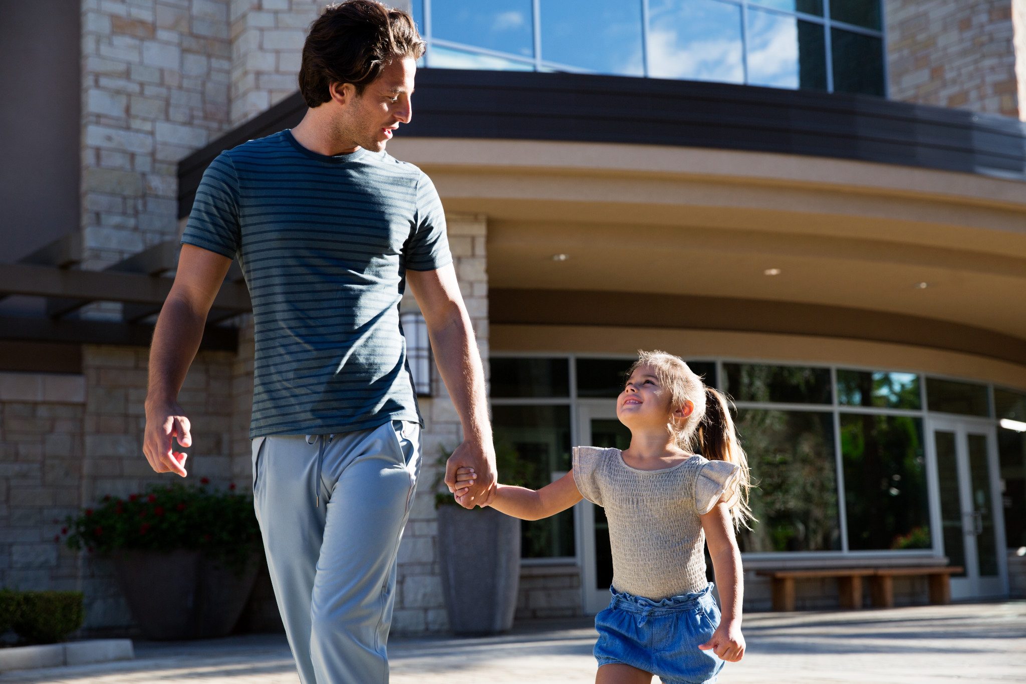 A man holds a female child’s hand as they walk together outside a Life Time building