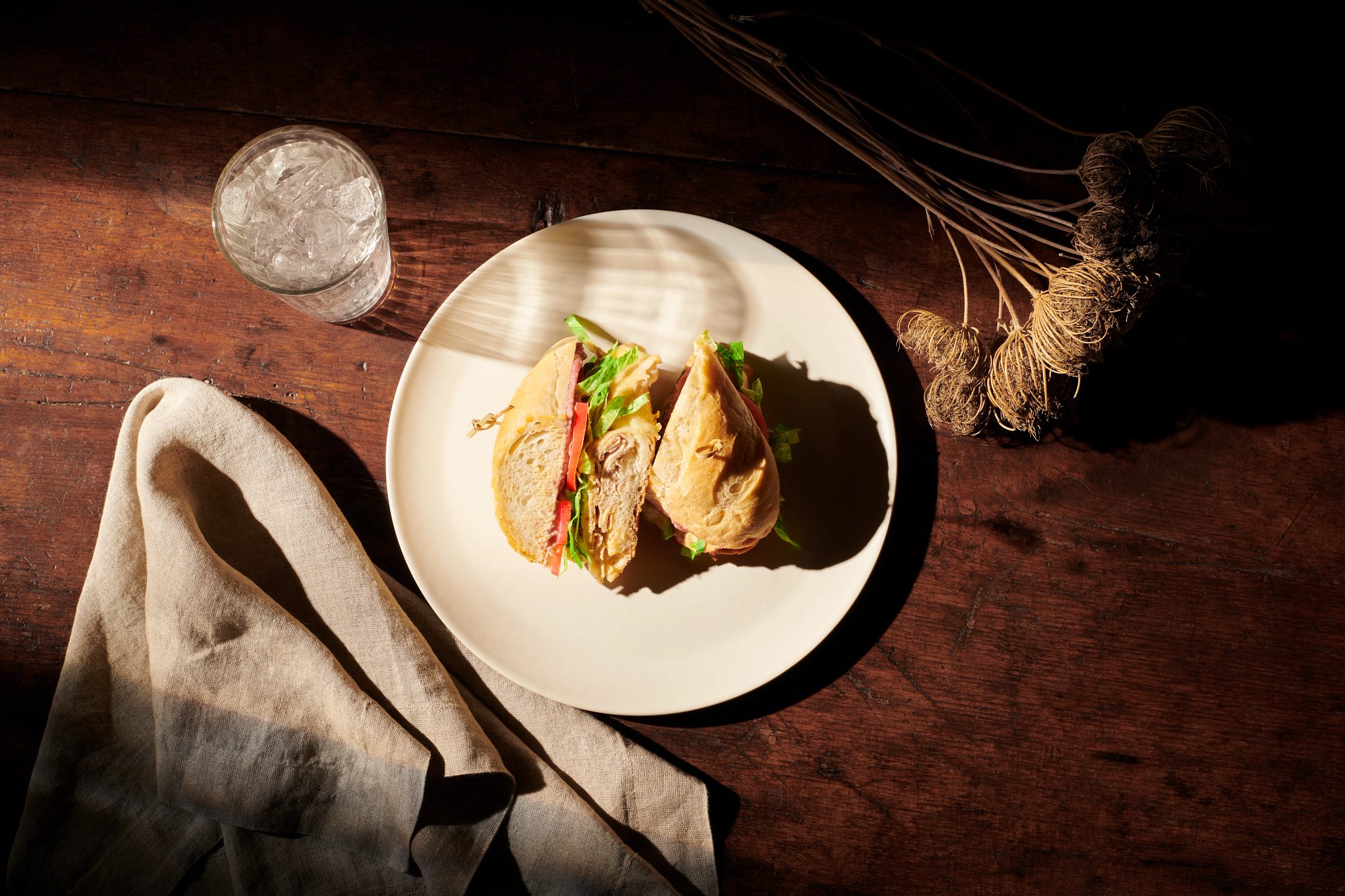 Overhead view of a turkey sandwich on a cream-colored plate placed on a dark wooden table with dried flowers and a glass of water
