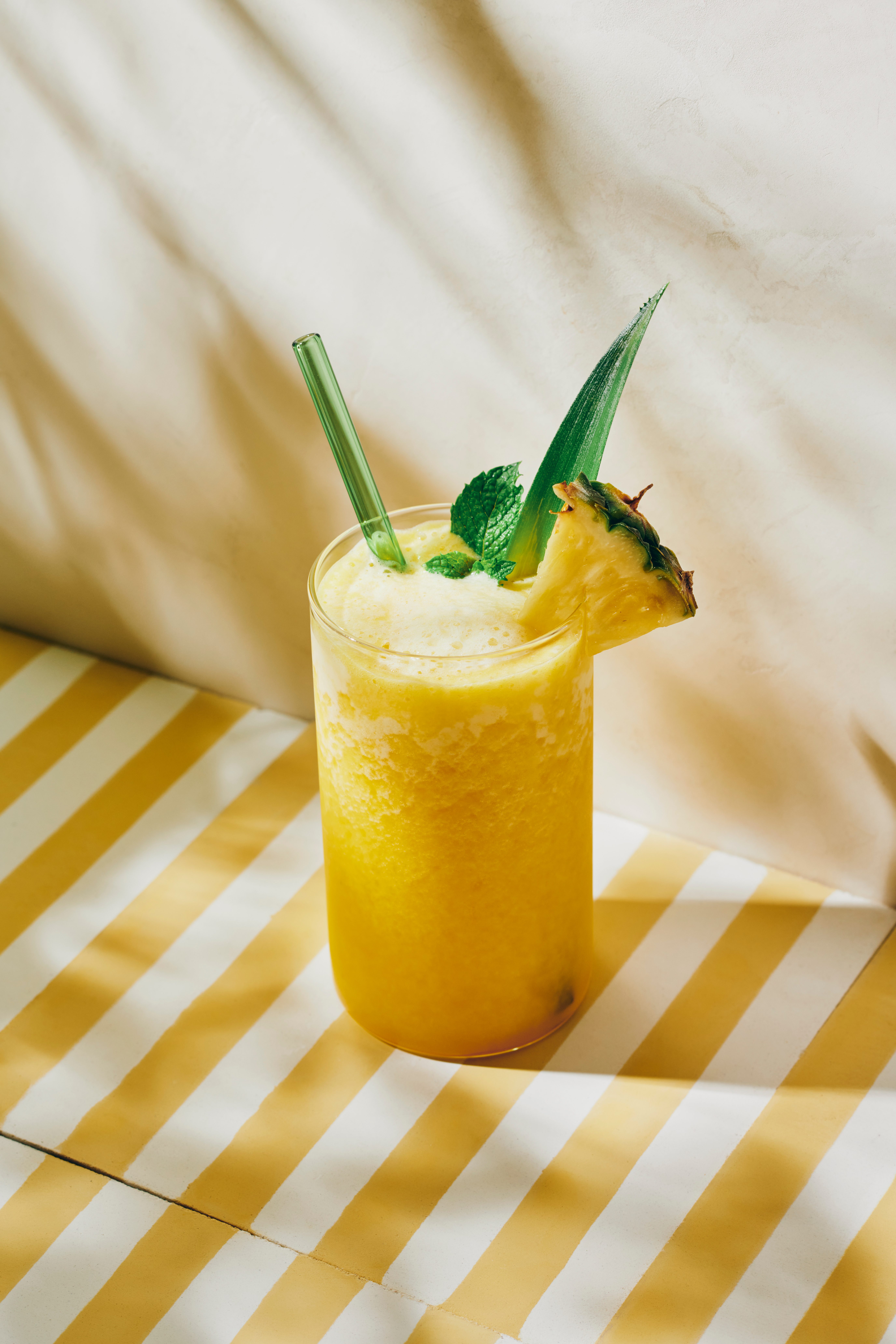 A pineapple smoothie garnished with a fresh slice of pineapple and mint leaf placed on a yellow and white striped tabletop
