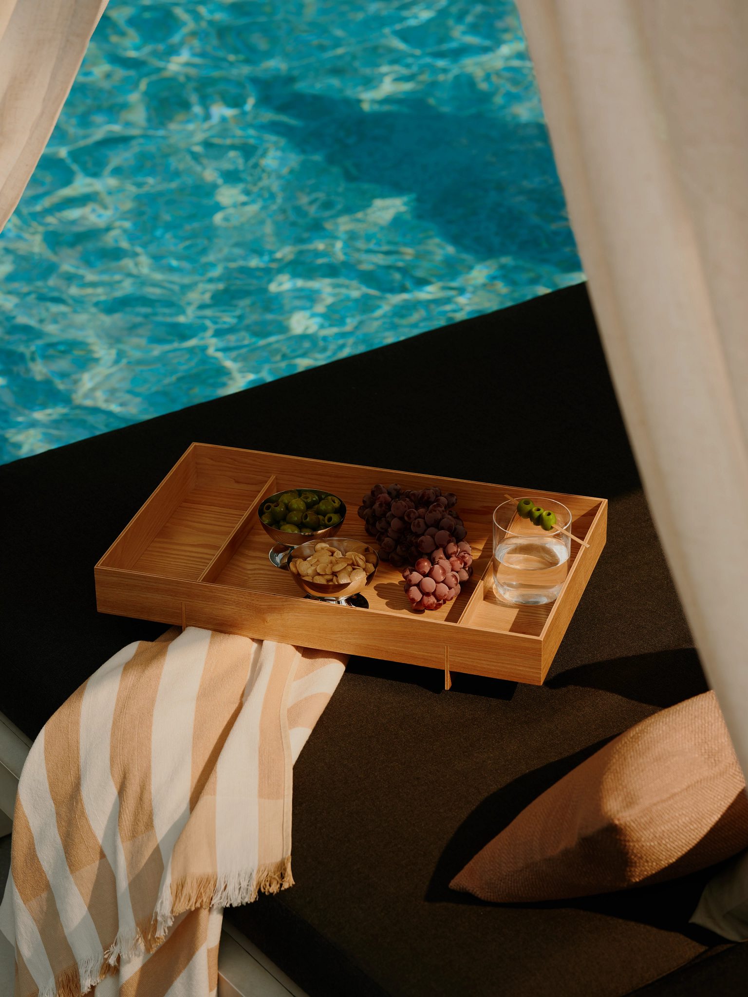 A wooden tray with food and drink atop a poolside cabana lounger
