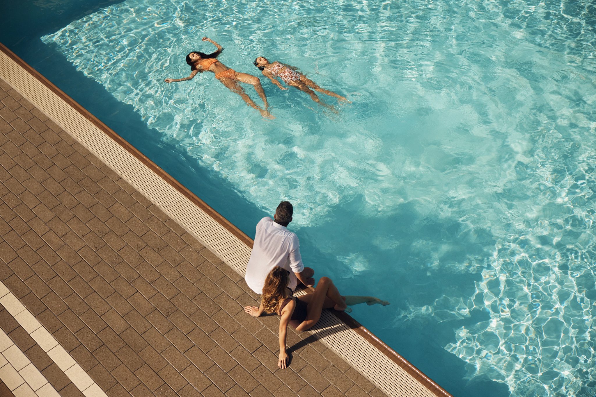 Mother and father sitting on the edge of an outdoor pool watching their children swim
