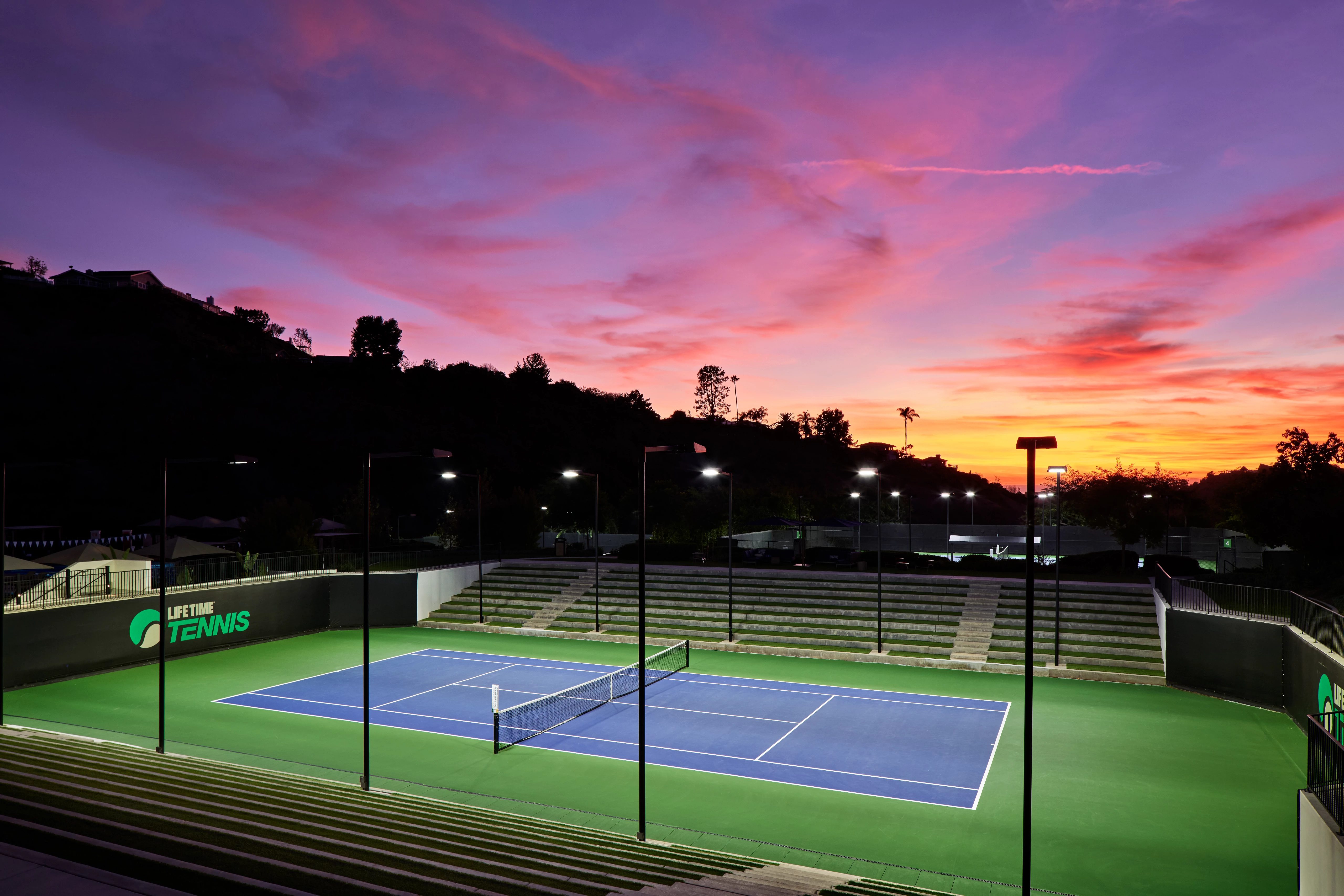 An outdoor tennis court lit by lights in the evening, dramatic colorful sky in the background