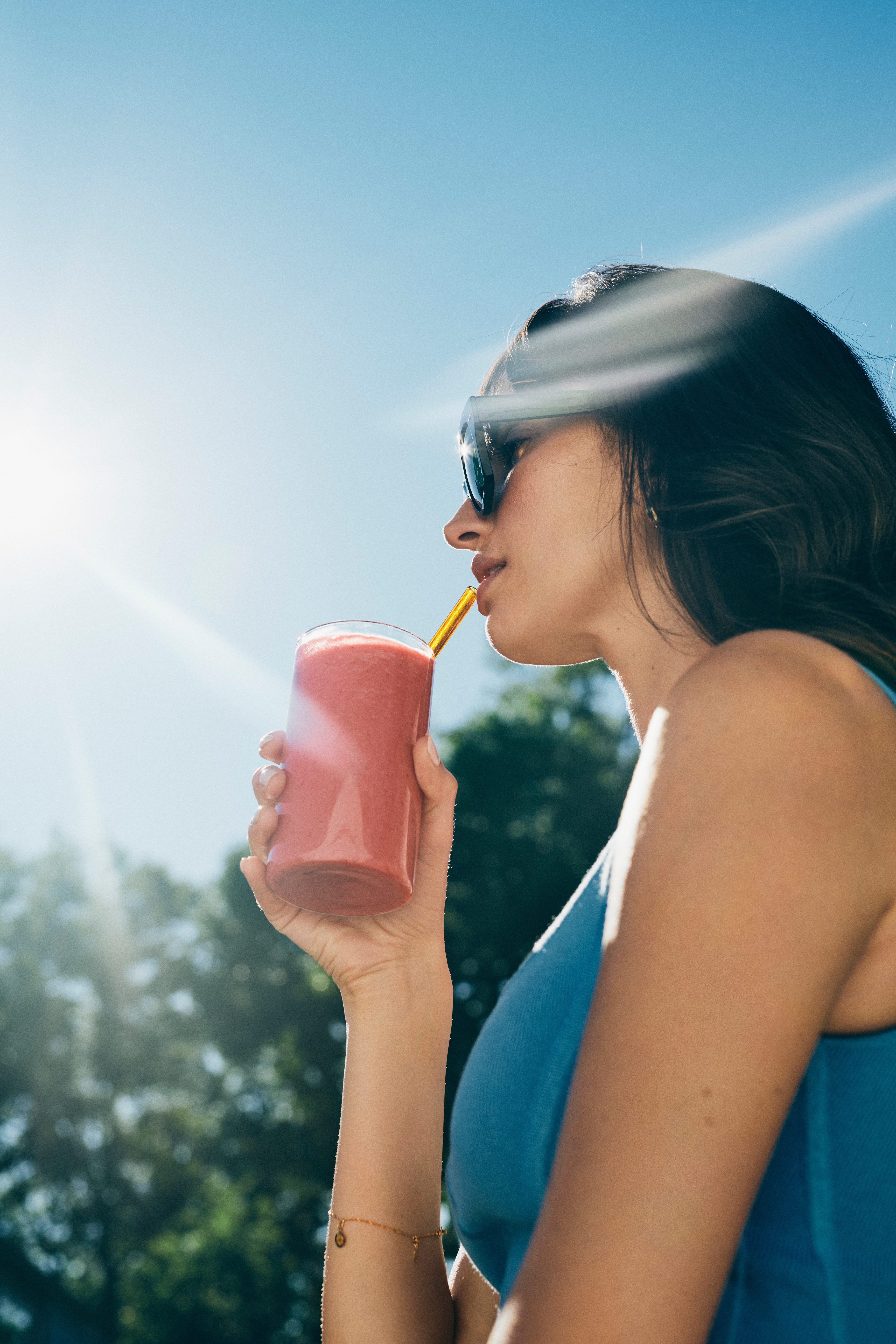 An attractive female wearing oversized sunglasses on a sunny day drinking a pink-colored smoothie