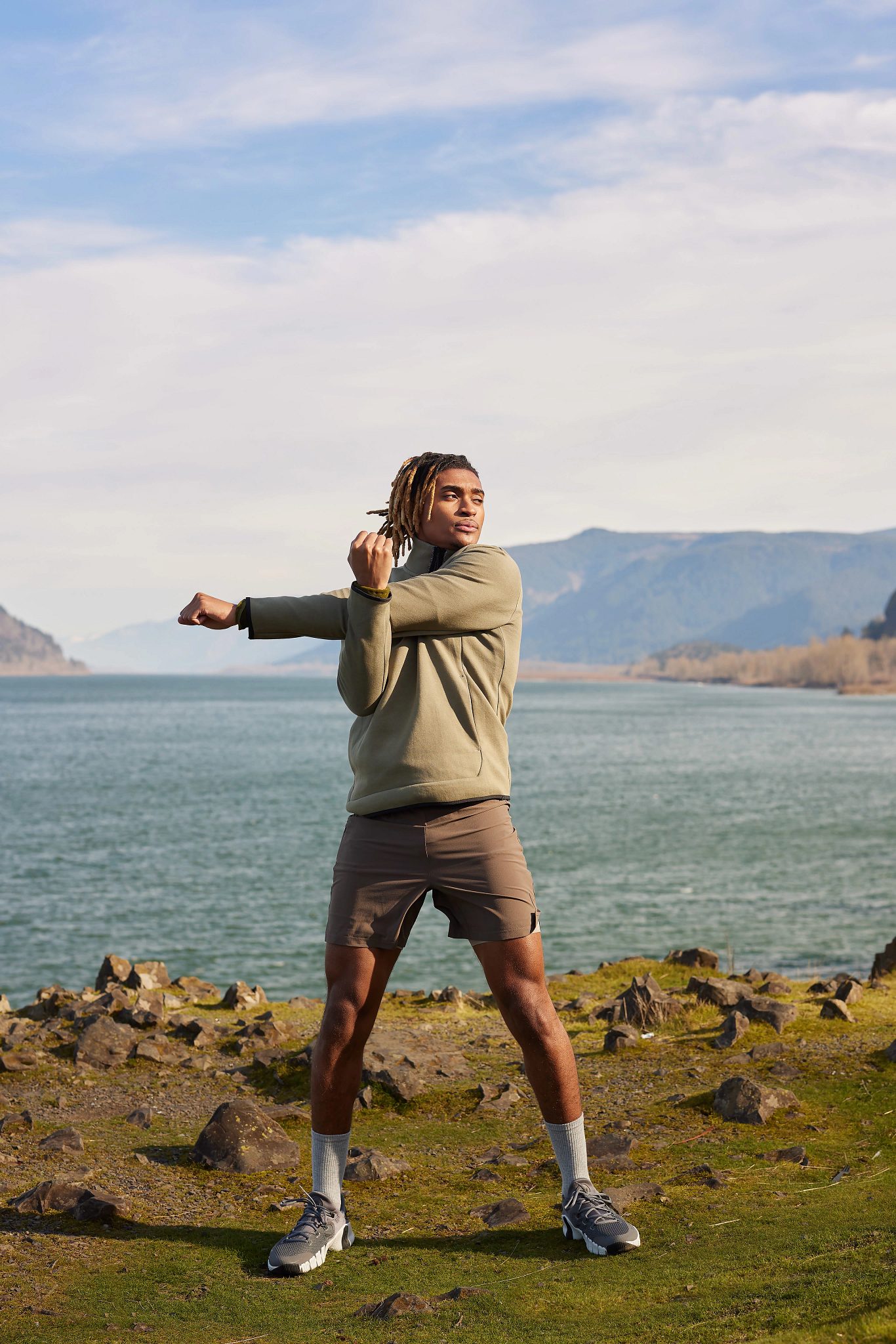 An adult male, standing next to water with mountains in the background, stretching his arms