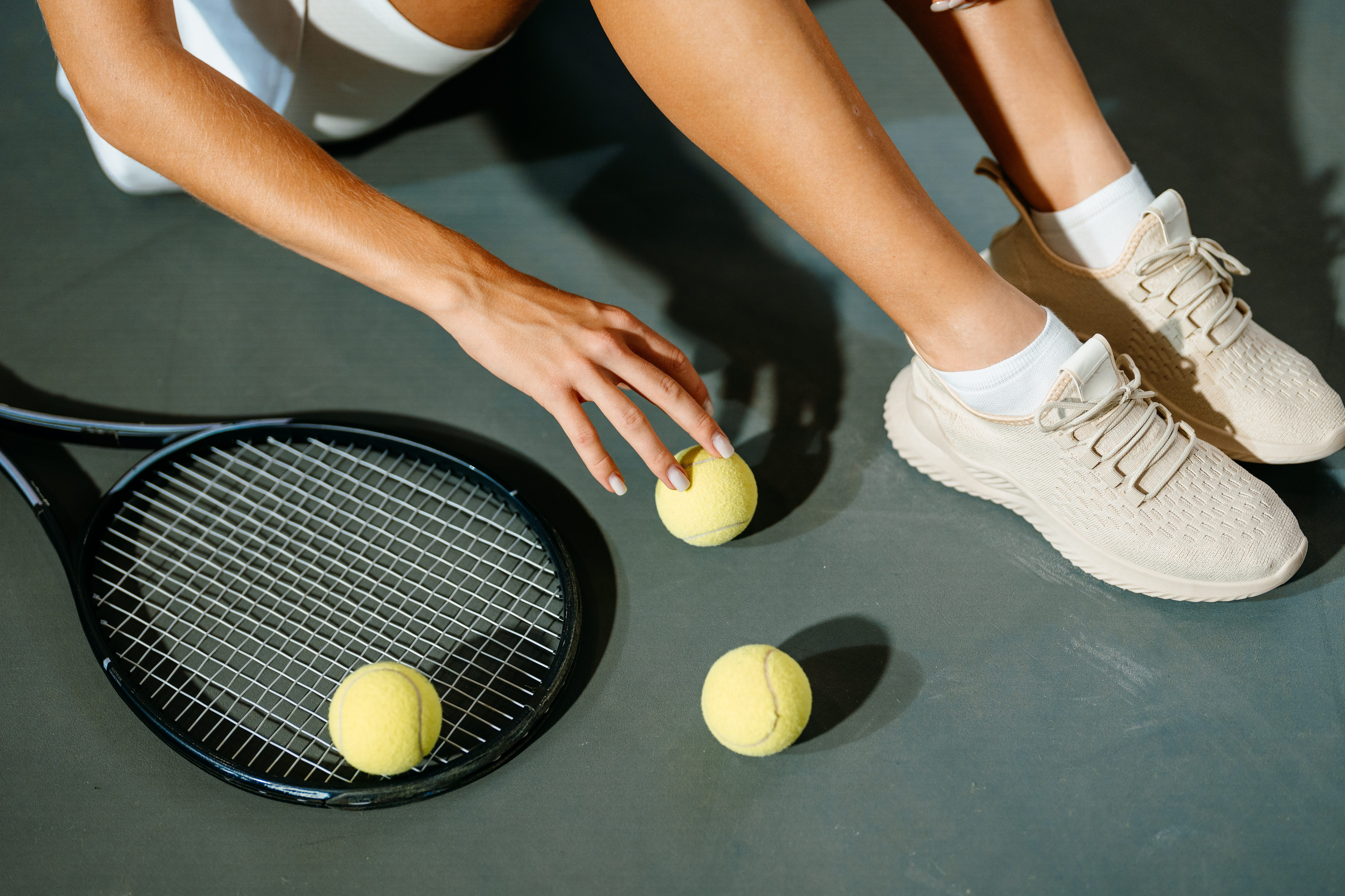 A woman in sportswear is sitting on the floor of a tennis court. A tennis racket and tennis balls are lying on the floor.