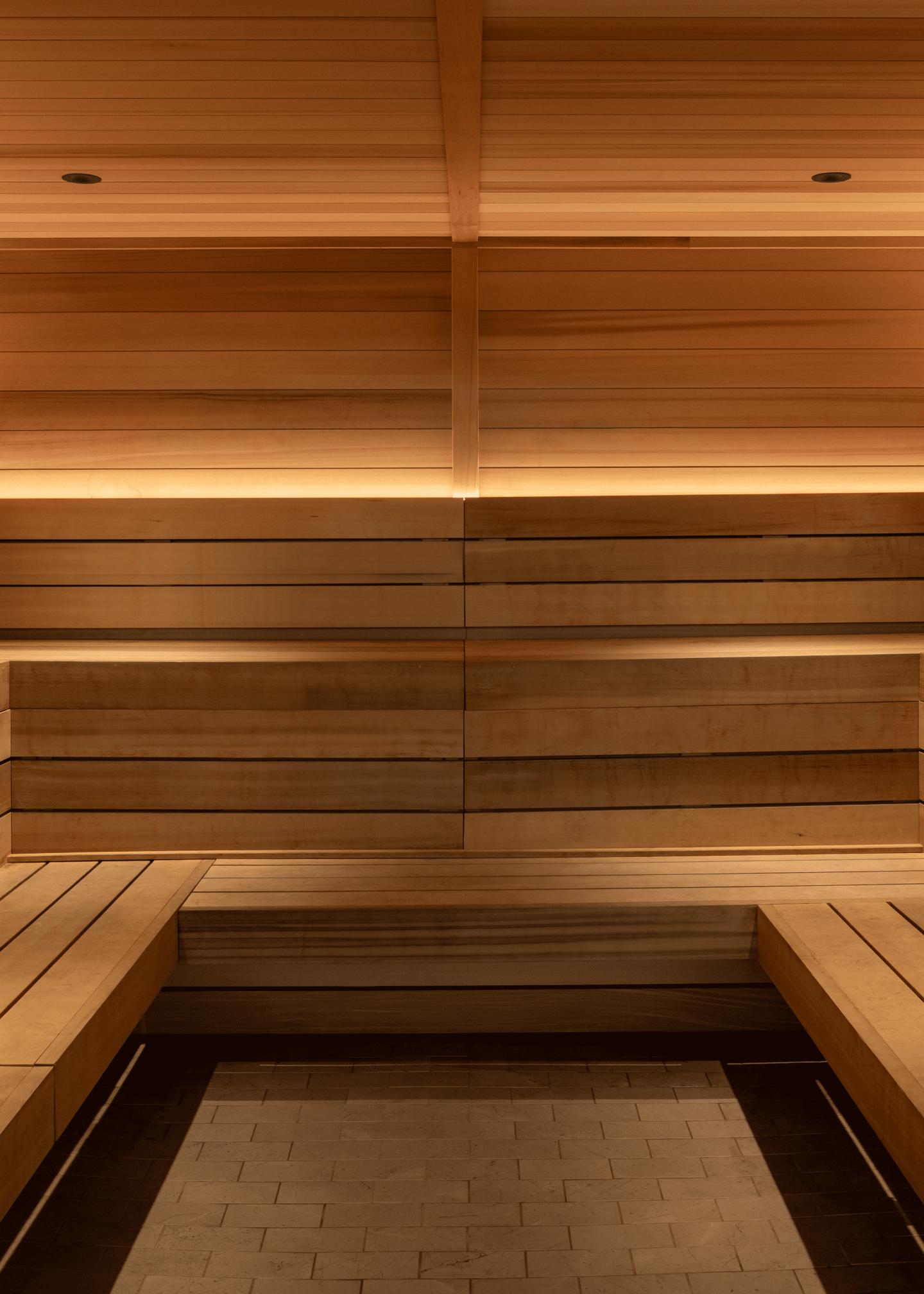 Modern wooden sauna with built-in benches on both sides, warm ambient lighting, and a stack of folded white towels on one of the benches.
