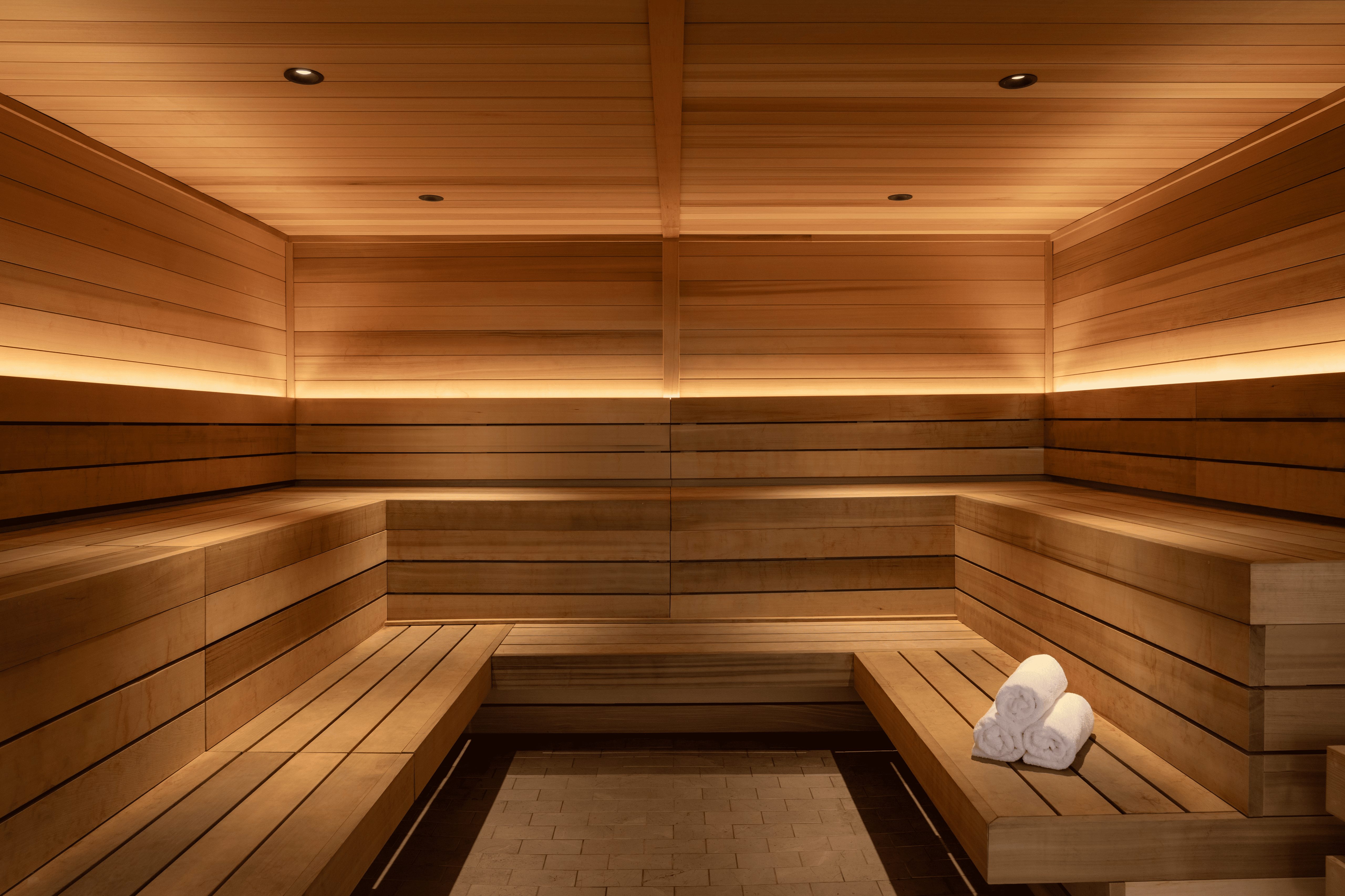 Modern wooden sauna with built-in benches on both sides, warm ambient lighting, and a stack of folded white towels on one of the benches.