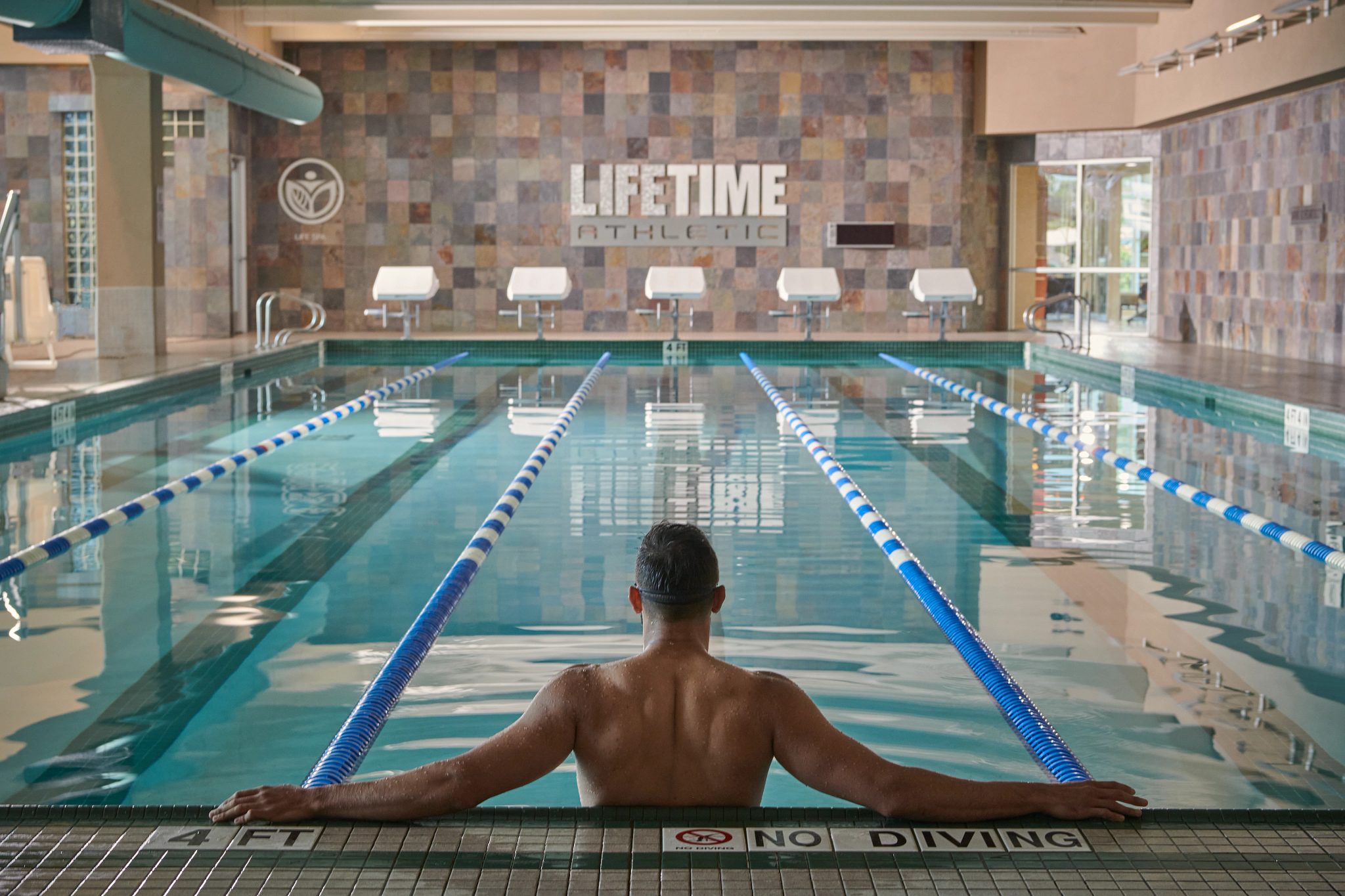 A man standing in an indoor lap pool; back to the camera