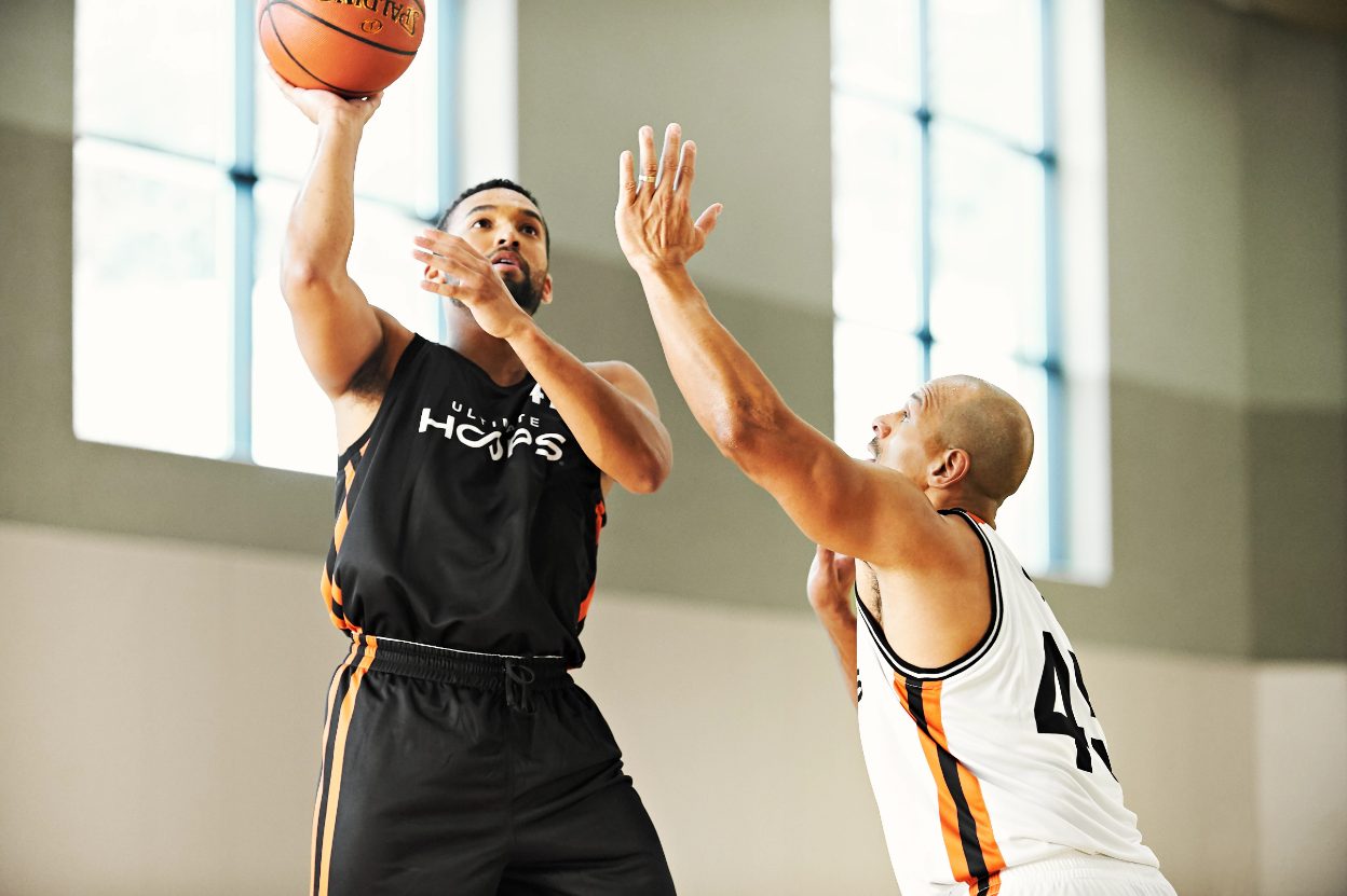 two men engaged in a basketball game inside a gym, dribbling and preparing to shoot at the hoop.