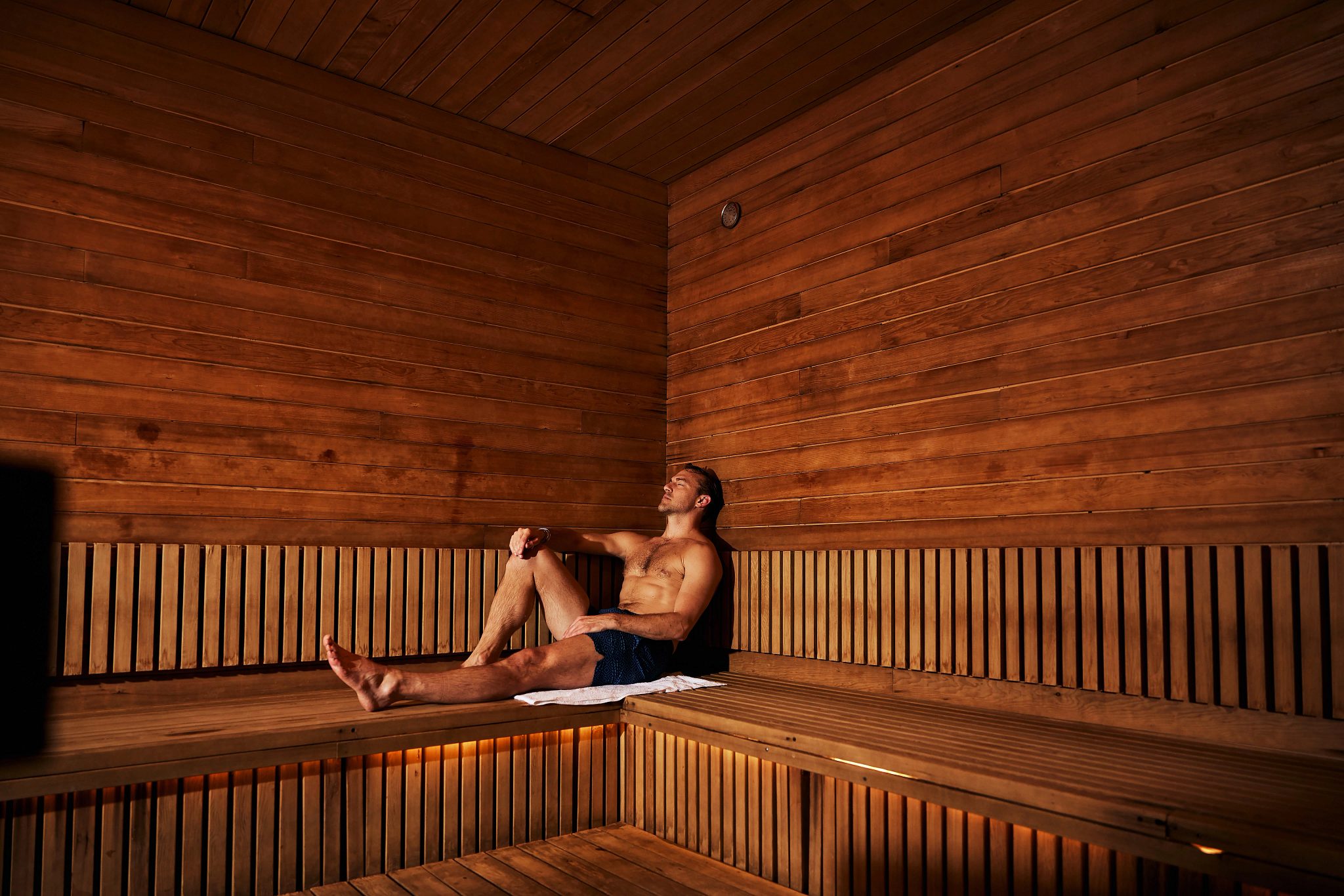 Man sitting in a sauna with his legs out and leaning against the wall with his eyes closed.