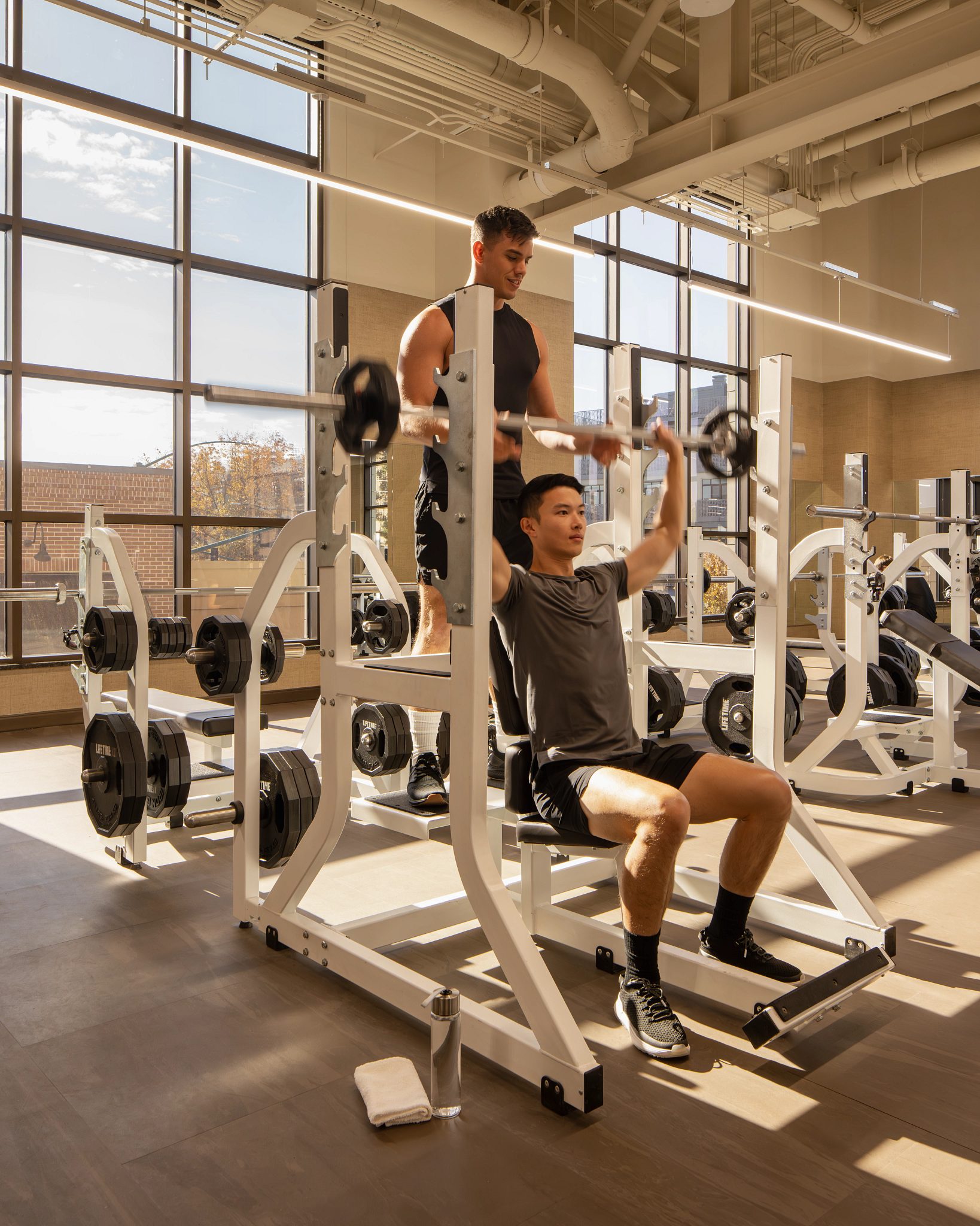 A man spotting another member while he completes overhead presses while seated on a weight bench