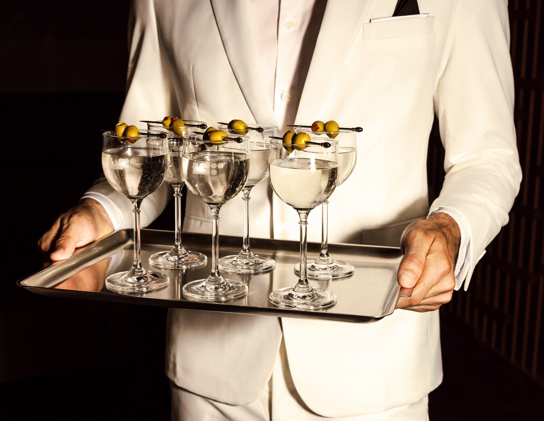 Professional bartender presenting craft cocktails - elegant server in white suit holding tray of premium drinks with olive garnish.
