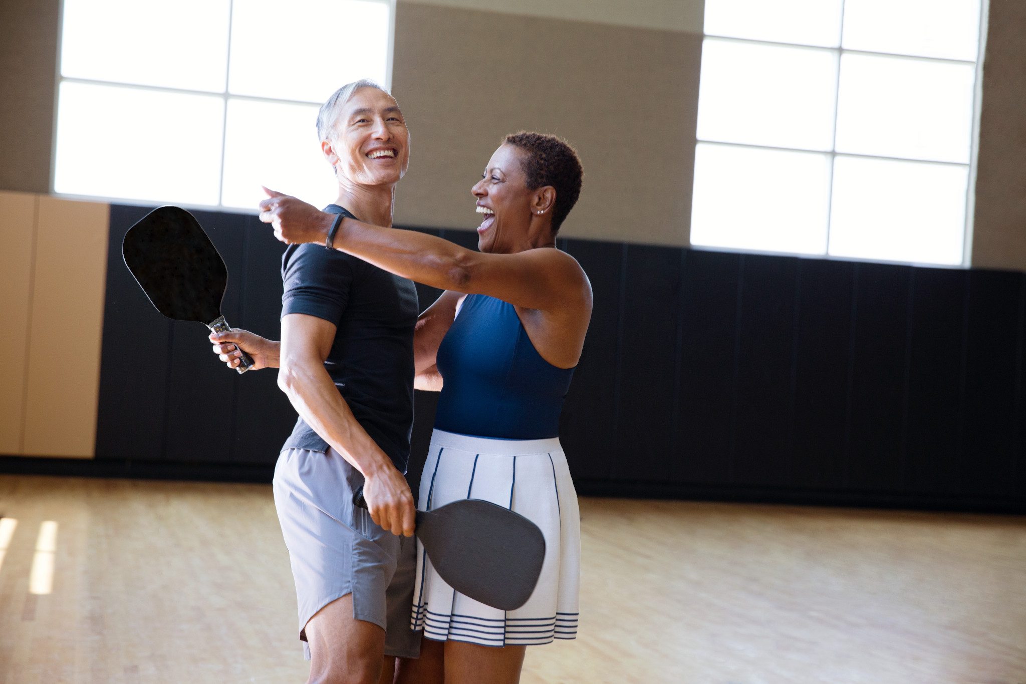 A man and a woman each holding a pickleball racuqet about to embrace eachother while standing on an indoor pickleball court