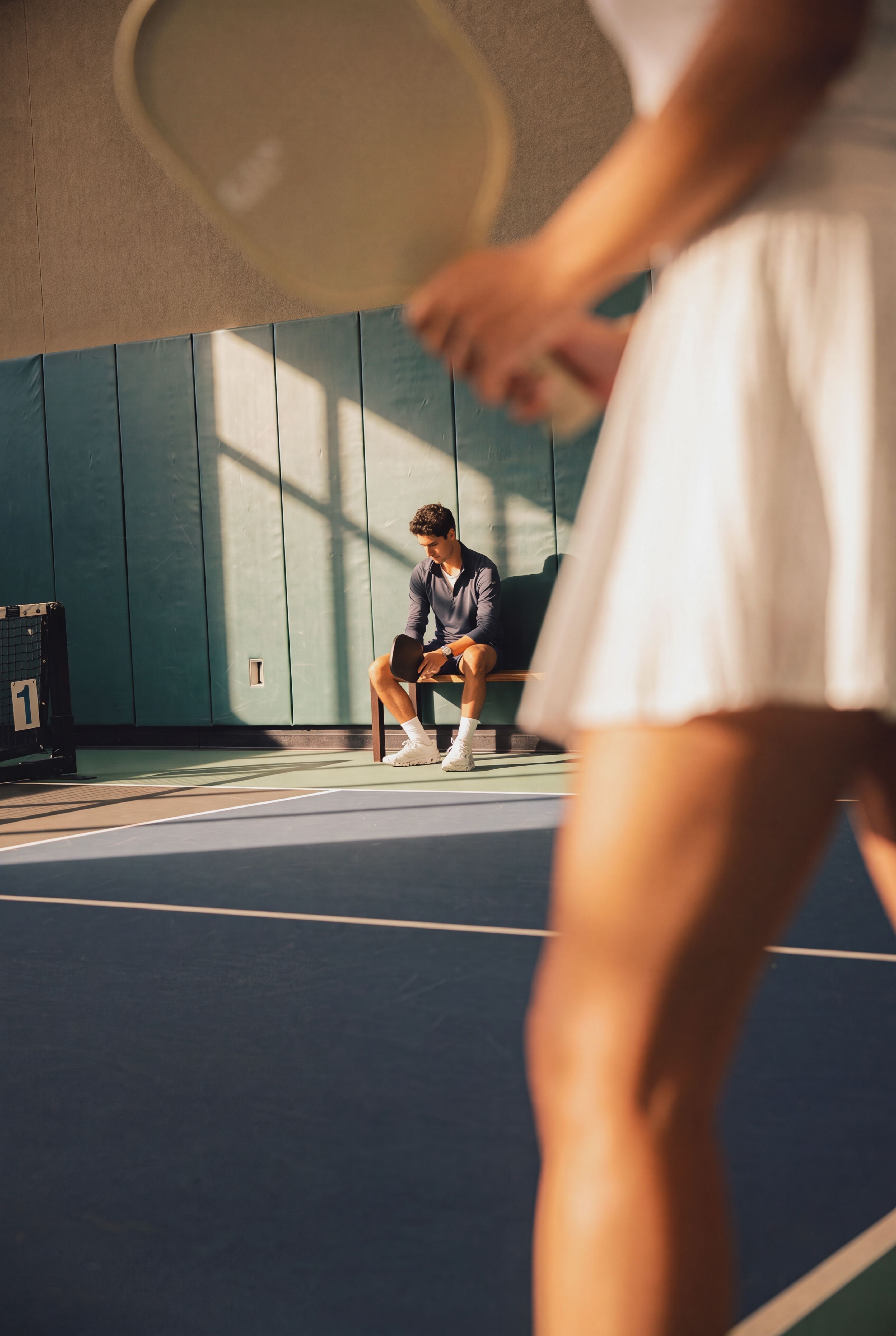 Extreme closeup of a woman on an indoor pickleball court holding a paddle, man sitting on a bench on the sidelines
