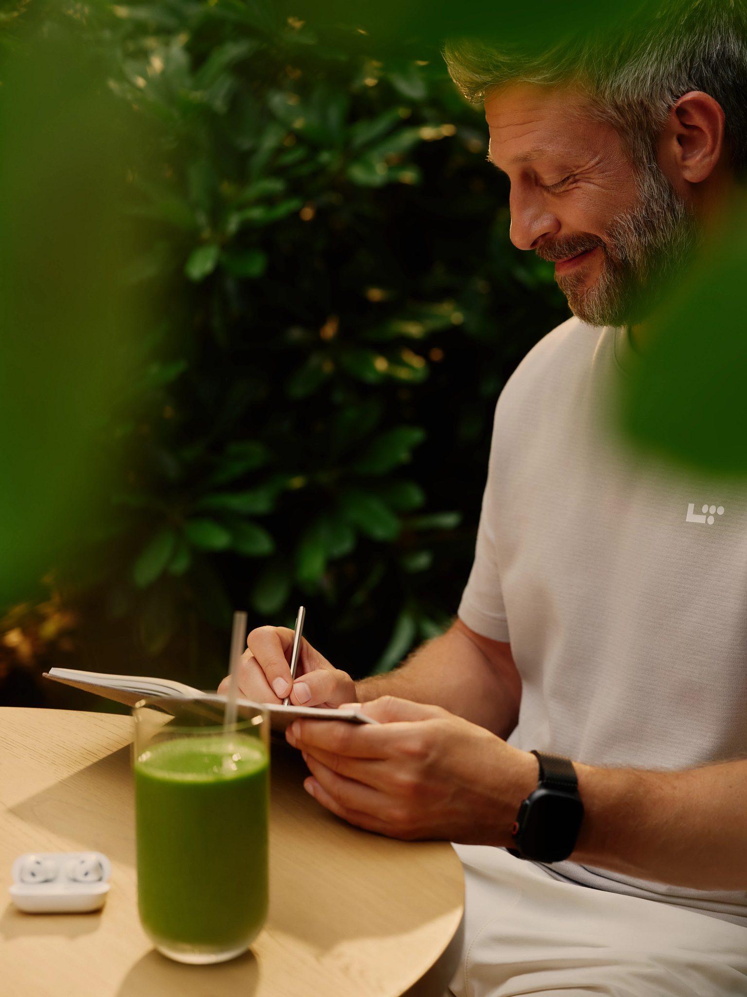 A man sitting outside writing in a journal with earbuds and a glass of green juice sitting on the table next to him