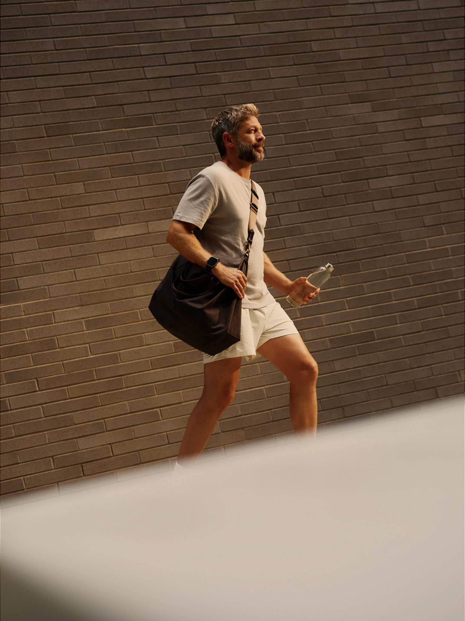 A man walking while carrying a glass water bottle and a gym bag; brick wall in background